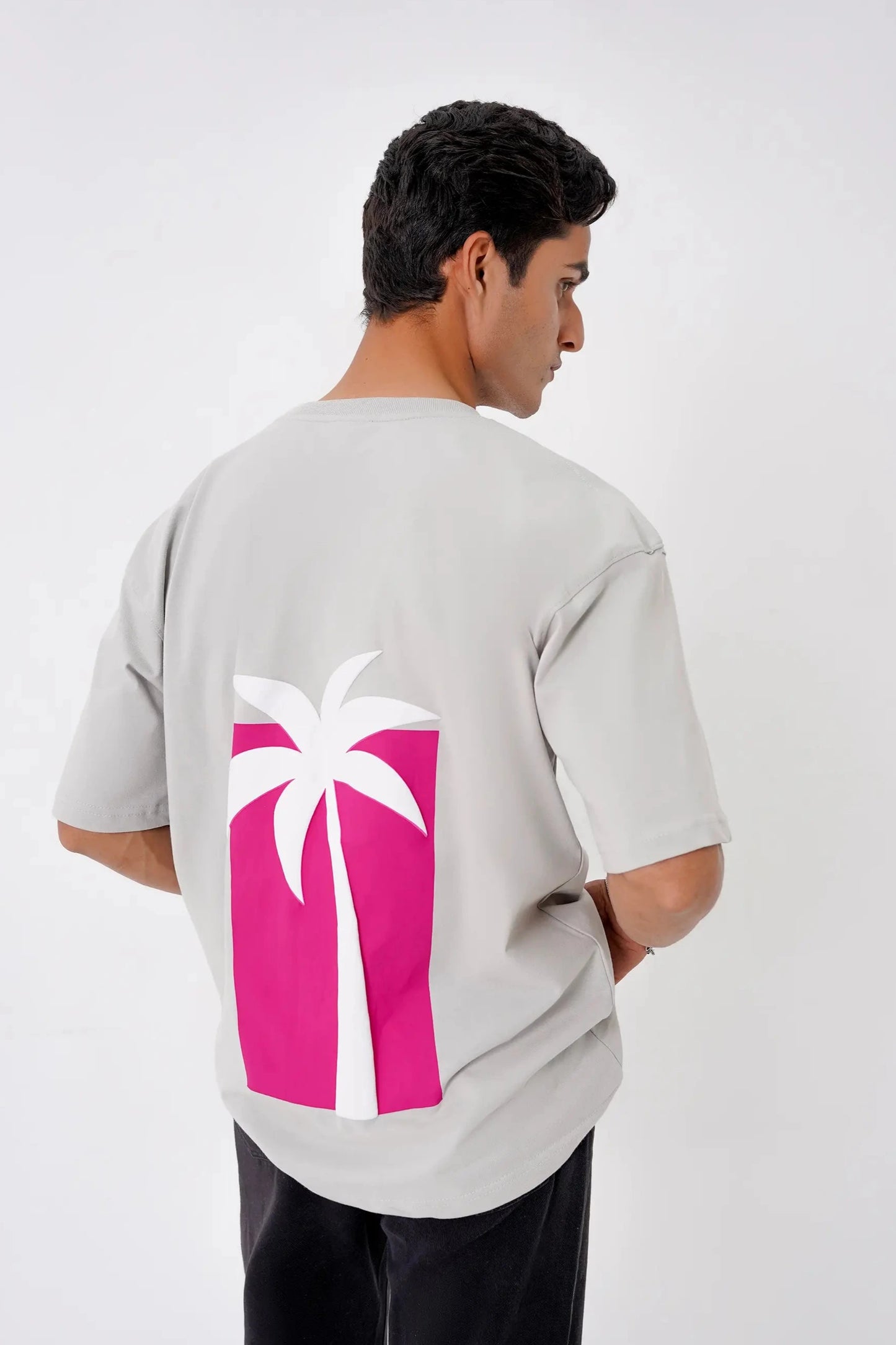Person wearing a light gray t-shirt with a pink and white palm tree design on the back, standing against a white background.