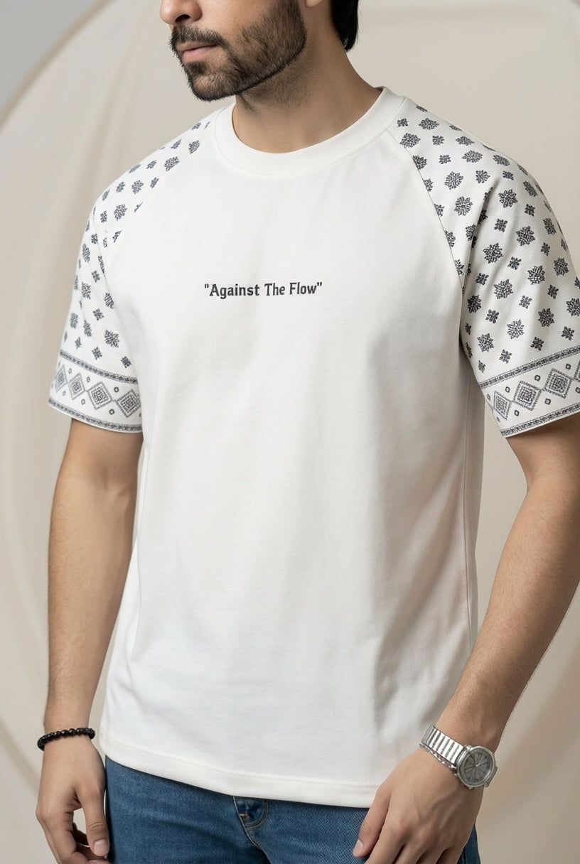 A man wearing a white short-sleeve t-shirt with black geometric patterns on the sleeves and the text "Against The Flow" printed on the chest, paired with blue jeans.