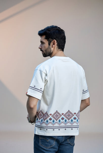 A man shown from the back wearing a white short-sleeve t-shirt with geometric embroidery in blue, purple, and red along the hem and sleeve cuffs, paired with blue jeans.
